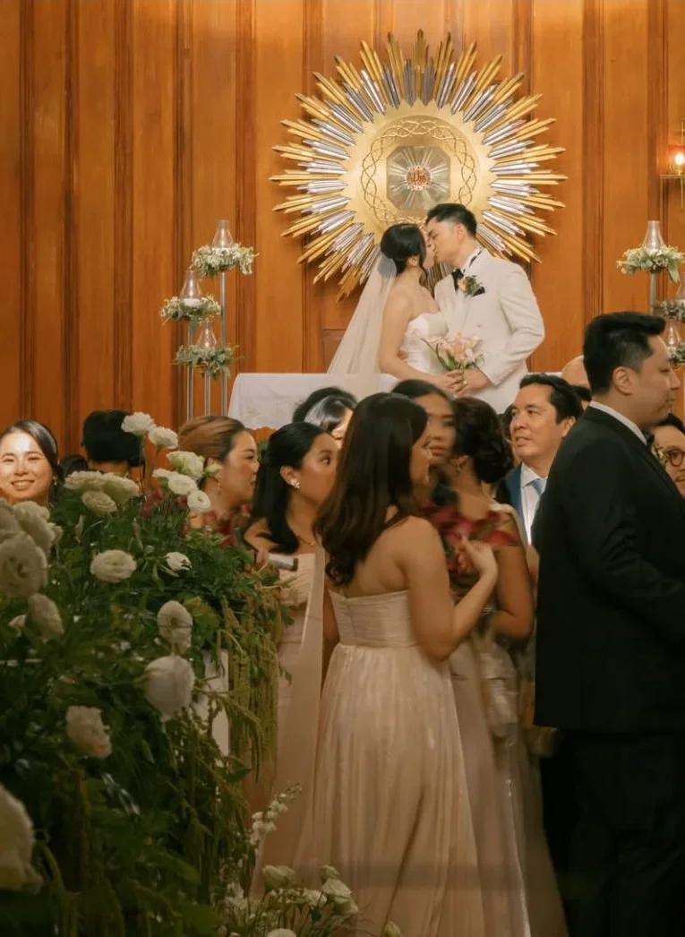 A couple kisses at the front of a wood-paneled room, with guests in the foreground.