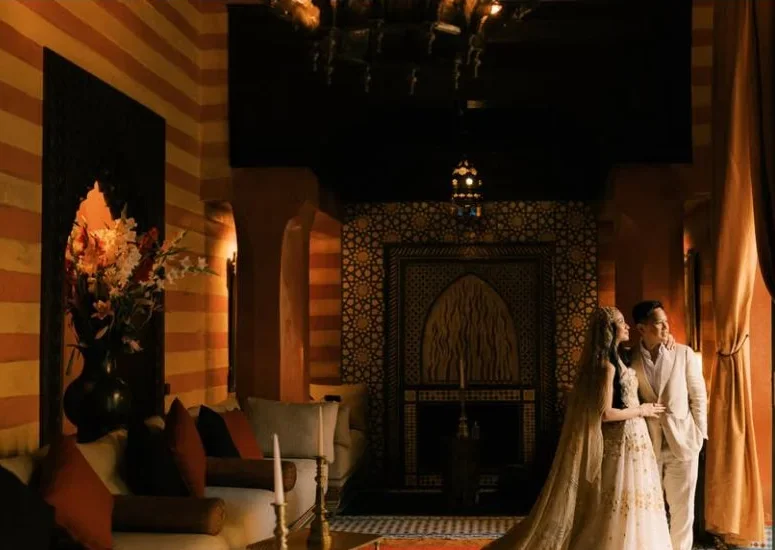 A couple beside a sunlit window in a richly patterned interior.