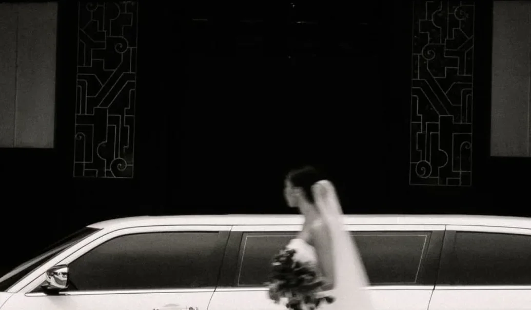 Black-and-white photograph of a bride beside a white limousine.