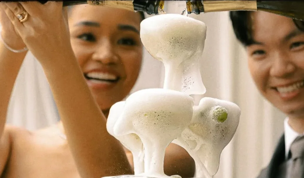 A couple smiles behind an overflowing champagne tower.