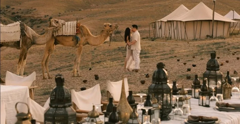 A couple beside camels and a tent in a desert setting.